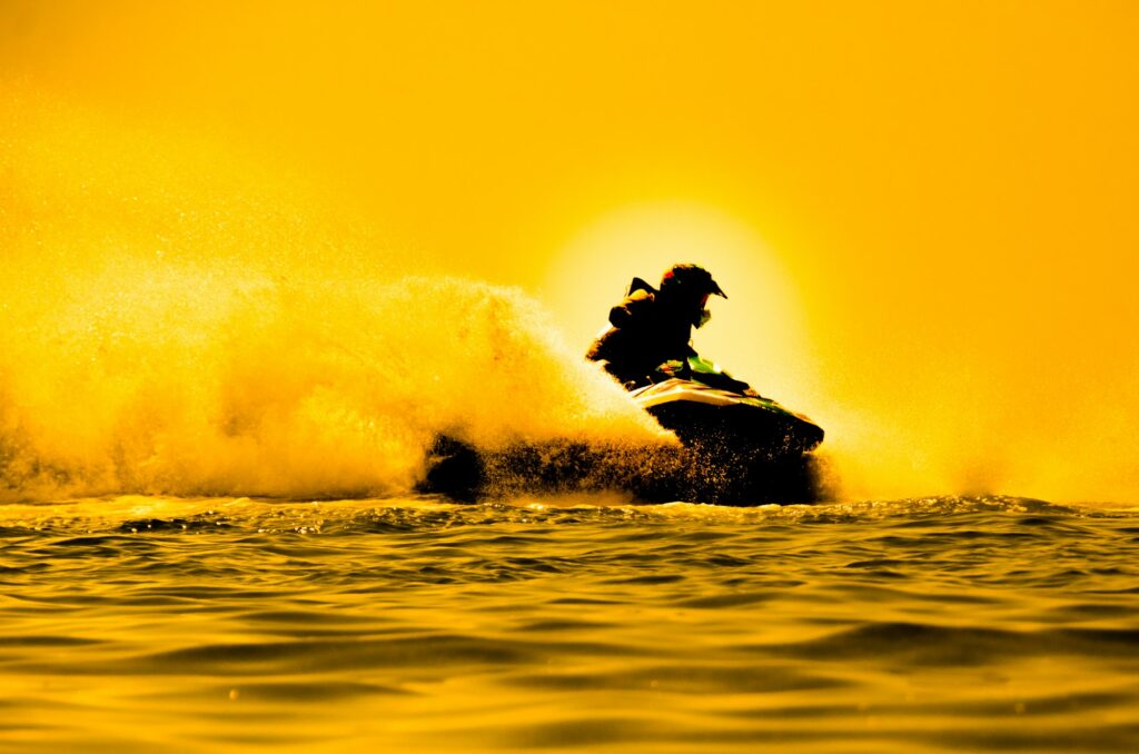 Silhouette of a person riding a jet ski on water during sunset, creating a trail of spray behind. The golden light casts a warm hue over the scene, reminiscent of the smooth finish achieved through paint protection film training.