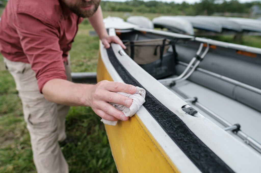 A person in a red shirt is meticulously cleaning the edge of a yellow kayak with a cloth on a grassy area, showcasing their skills as if undergoing paint protection film training.