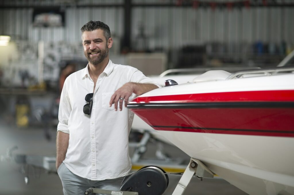 A man wearing a white shirt and sunglasses leans against a red and white boat, amidst the industrial backdrop of a warehouse, looking like he could be waiting to start Paint Protection Film Training.