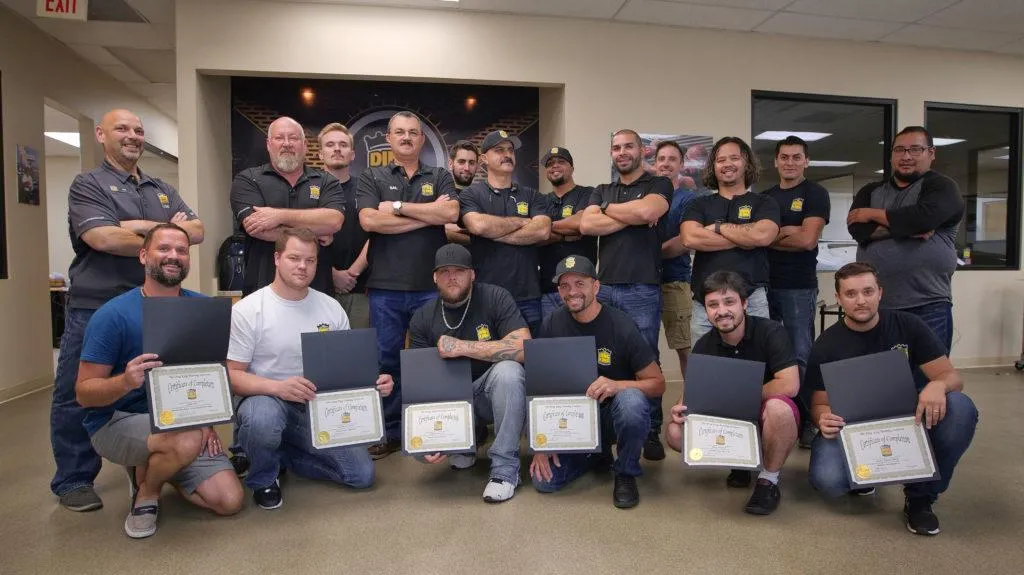 A group of men in a room, some standing with folded arms and others kneeling, proudly displaying certificates and folders from their Paint Protection Film Training.