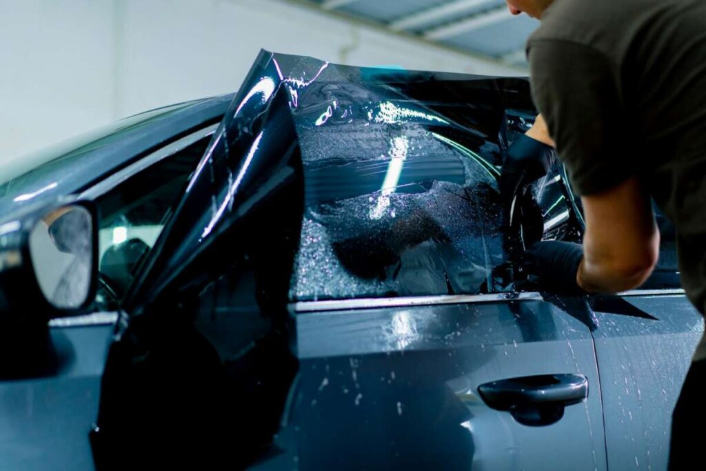 In a garage setting, a person skillfully applies window tint and PPF to a car door with sleek black film, enhancing both privacy and windshield protection.