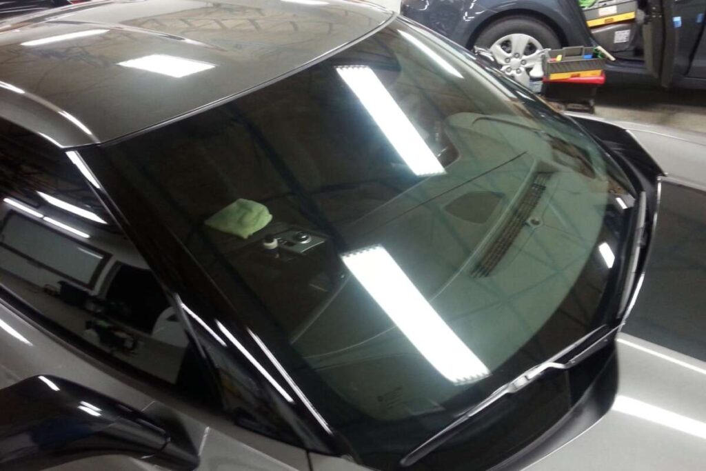 A silver car with a pristine windshield reflects the overhead lights in a garage setting, hinting at the owner's dedication from recent paint protection film training. A cleaning cloth rests on the dashboard, completing the sleek, polished look.