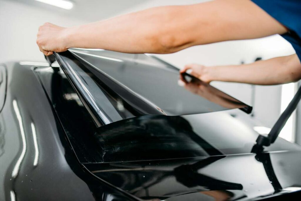 In a well-lit workshop, a person carefully applies ceramic window tint film to a car window, ensuring precision and quality.