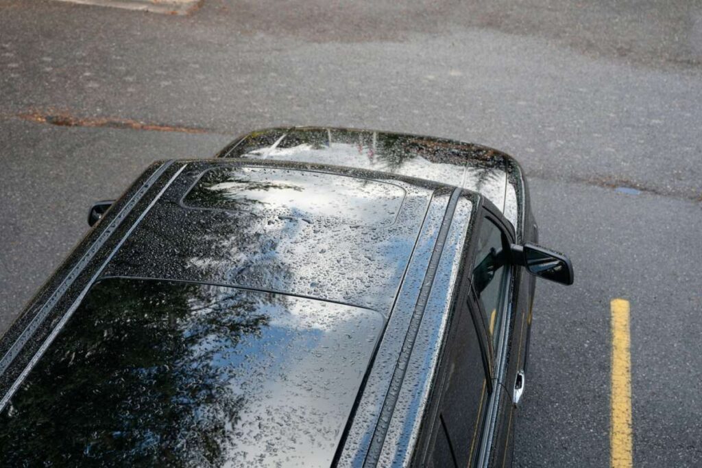 Top view of a black car with raindrops glistening on its roof, showcasing its sleek PPF finish, parked beside a yellow line on the wet asphalt surface.