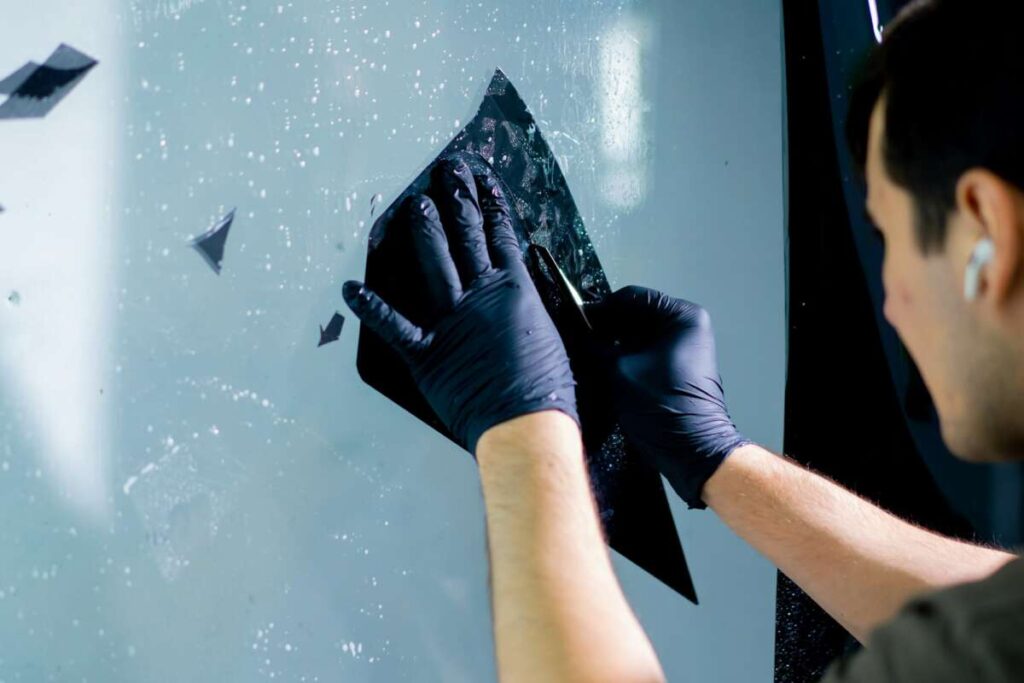 A person wearing gloves carefully applies the window tint film to a glass surface, ensuring it aligns perfectly for optimal protection, much like precision seen in PPF windshield protection film applications.