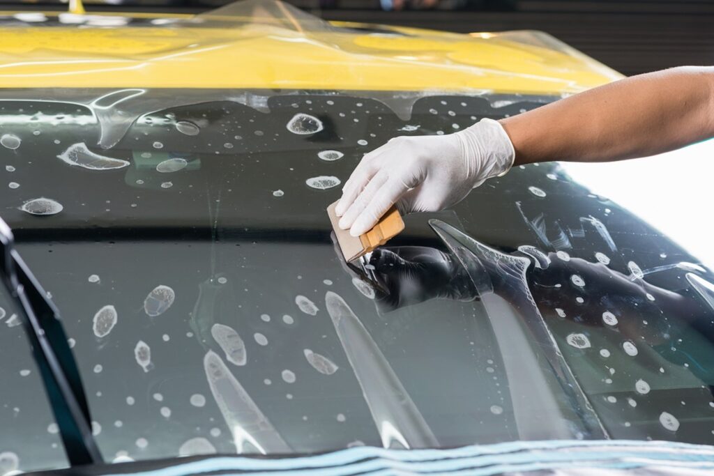 A gloved hand applies film to a car windshield using a squeegee tool, expertly smoothing out air bubbles during Paint Protection Film Training.