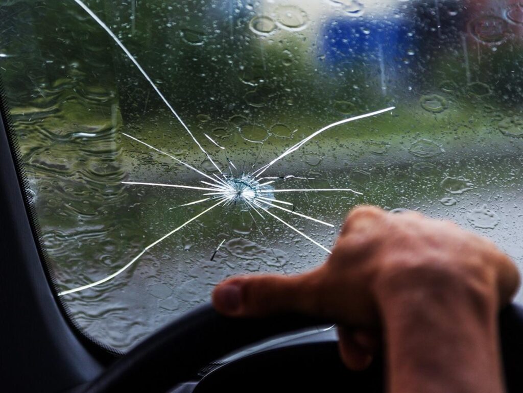 Close-up view of a cracked windshield on a rainy day, with a person's hand on the steering wheel, reflecting the importance of proper paint protection film training for vehicle safety.