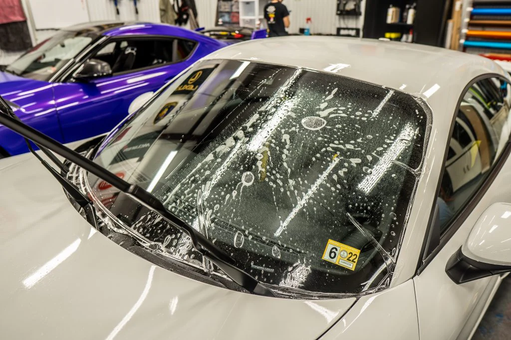 A white car with water and soap on its windshield in a garage, ready for Paint Protection Film Training, with its wiper blades lifted. A blue car is parked beside it.