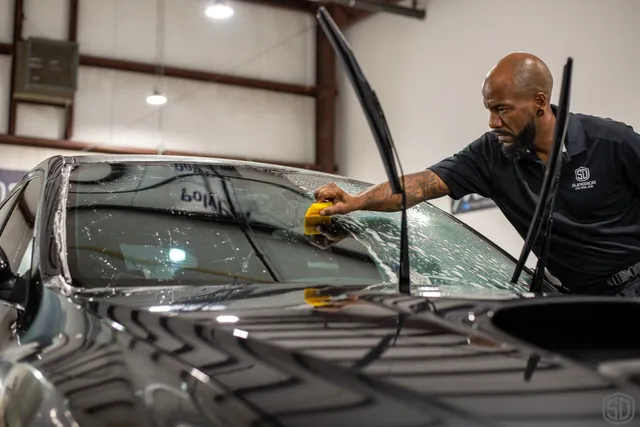 A person uses a squeegee to clean the windshield of a black car inside a garage, with the car's wipers raised, preparing it for Paint Protection Film Training.