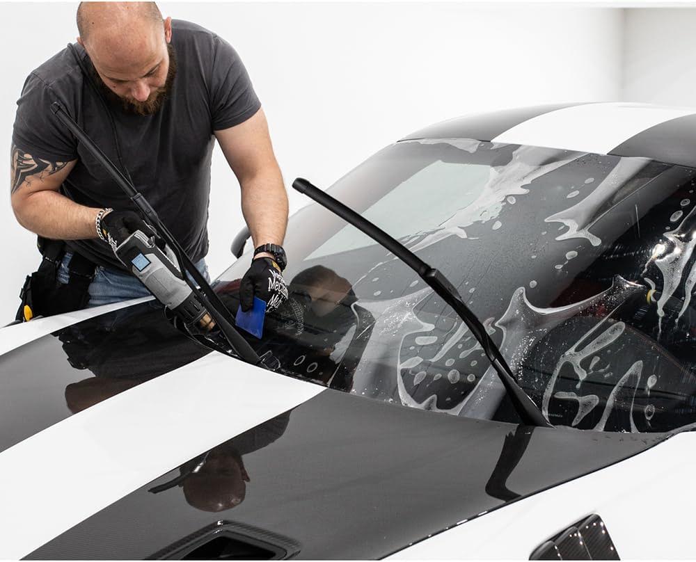 A man with a tattooed arm uses tools to apply a protective film to the windshield of a shiny black and white car, showcasing techniques from his Paint Protection Film Training.